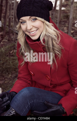 Weiblich, lange blonde Haare, trägt einen kurzen roten Zweireiher Mantel, schwarzen Hut, schwarze Handschuhe und Jeans, auf dem Boden sitzend, Stockfoto