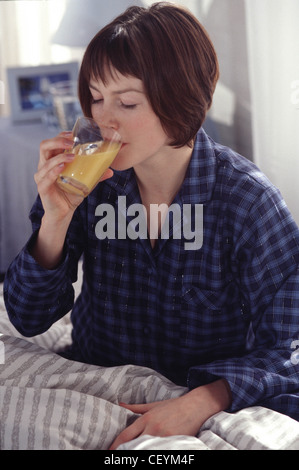 Weibchen mit kurze brünette Haare tragen blaue Tartan Pyjama sitzen im Bett trinken Orangensaft Stockfoto