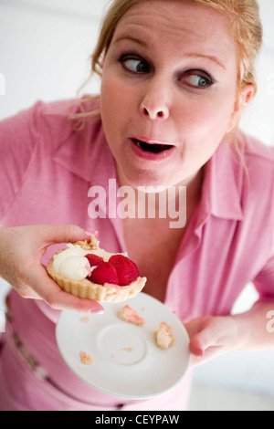 Nahaufnahme eines größeren weibliche blonde Haare aus Gesicht trägt rosa Hemd halten Platte und Erdbeer-Sahne-Torte in der hand, Mund Stockfoto