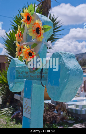 Ein Kranz aus künstlichen Blumen hängt an einem Kreuz geformten Grabstein auf einem Friedhof auf dem Quilotoa Loop in Ecuador. Stockfoto