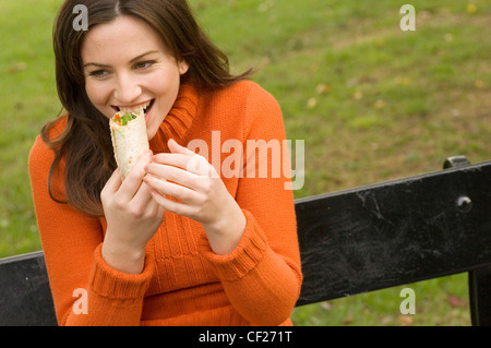 Frauen tragen orange Rollkragen Pullover sitzen auf der Parkbank Essen wrap Stockfoto