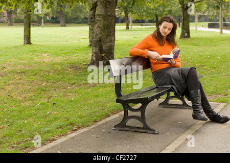 Weibchen tragen orange Rollkragen Pullover grau Rock und schwarze Stiefel sitzen auf der Parkbank Buch Stockfoto