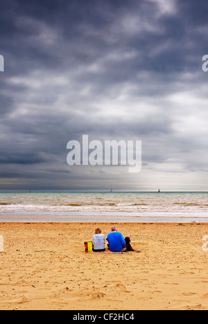 Ein paar sitzt mit ihrem Hund unter einem düsteren Himmel am Strand von Camber Sands. Stockfoto