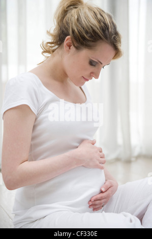 Schwangere Frauen blonde Haare gebunden zu Pferdeschwanz tragen ein weißes Hemd und weiße Hose, auf den Flowith Beinen sitzen Stockfoto