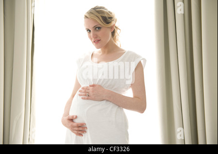 Schwangeres Frauen blondes Haar gebunden zu Pferdeschwanz, trägt ein weißes Kleid, stehend durch die Fenster Hände auf Beule, Lächeln, mit Blick auf Stockfoto