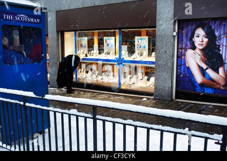 Eine Frau auf der Suche in der Auslage ein Juweliergeschäft gegenüber einen Zeitungskiosk, während der Schnee in der Princes Street fällt. Stockfoto