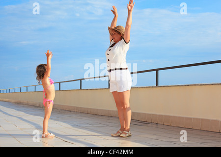 ältere Frau und kleine Mädchen tun Frühsport auf Veranda in der Nähe von Seeküste, gehobenen Händen nach oben Stockfoto