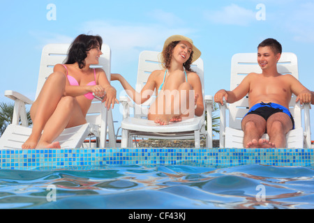 Lächelnd Mann und zwei junge Frauen lehnen sich auf Liegesessel in der Nähe von Open-Air-Pool, miteinander reden Stockfoto