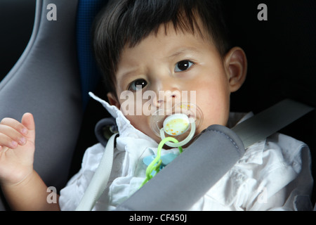 Cute Baby eurasischen Boy in einem Baby-Sitz-Auto hat seinen Schnuller in den Mund. Er schaut uns an. Es ist eine Nahaufnahme zu schießen. Er trägt ein weißes Stockfoto