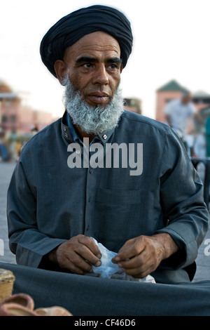 Einem lokalen Verkäufer in Tracht, Keffiyeh Schal Kopfund Bart sitzt auf seinen Platz innerhalb der Djemaa el-Fna Markt Ma verkaufen Stockfoto