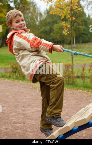 Junge in Jacke ist auf Spielplatz im Herbst Park. Lächelnd, steigt er die Treppe an einem Seil festhalten. Stockfoto