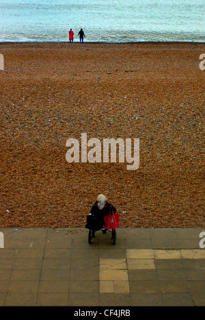 Eine ältere Frau in einem Rollstuhl von Brighton Beach mit Blick auf ein junges Paar in der Ferne. Stockfoto