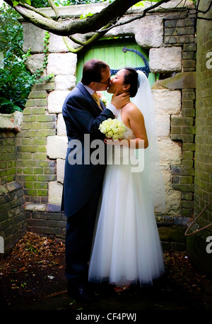 Hochzeitspaar sind unter Baum küssen. Ein Bräutigam hält Braut Hals und küsste sie auf die Lippen. Braut hält Blumen in Ihr Stockfoto