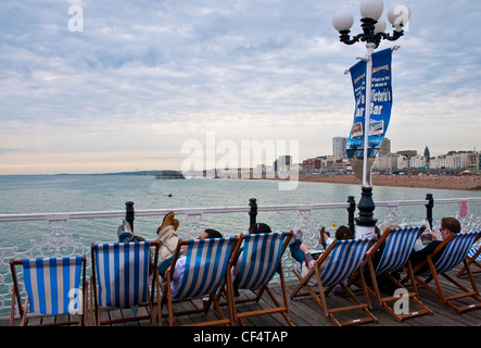 Menschen entspannen in Liegestühlen auf Brighton Pier mit Blick auf die Reste der West Pier. Stockfoto