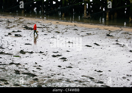 Ein Mann mit einer leuchtend roten Jacke ein Strand bei Ebbe, neben einem Holzsteg entlang spazieren. Stockfoto