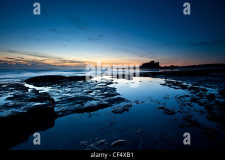 Blick über das felsige Ufer in Richtung Bamburgh Castle Silhouette von frühen Dämmerlicht. Stockfoto