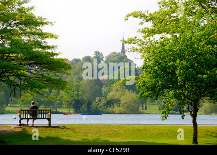 Ein Mann sitzt auf einer Parkbank Blick über einen See in Wimbledon Park. Stockfoto