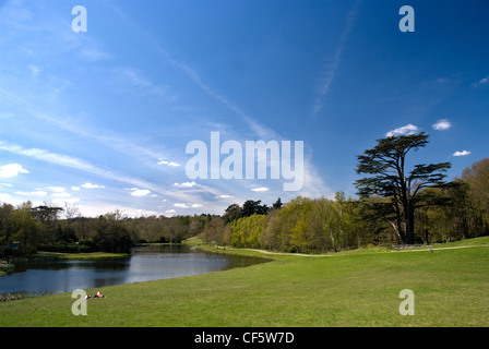 Ein paar entspannende auf dem Rasen an einem See im Painshill Park, einem sorgfältig restaurierten 18. Jahrhundert Landschaft Park entworfen und erstellen Stockfoto