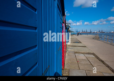 Ein paar gehen arm in Arm vorbei vor kurzem bemalte hölzerne Strandhütten. Felixstowe Docks sind im Hintergrund. Stockfoto