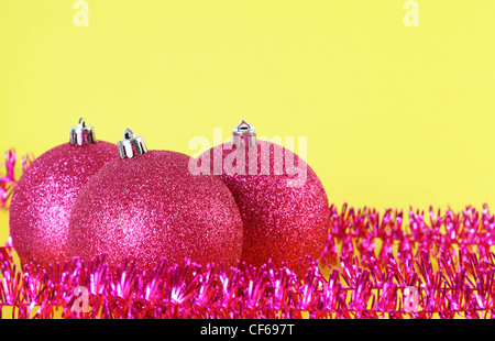 Oben drei rote Christbaumkugeln umgeben von lila Lametta isoliert auf gelb, vordere Kugel im Fokus. Stockfoto