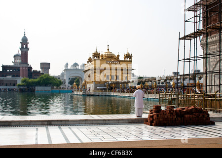 Beten am Rand des Teiches im Inneren des goldenen Tempels aus Sicht der Amrit Sarovar, Darbar Sahib und Damm über dem Wasser Stockfoto