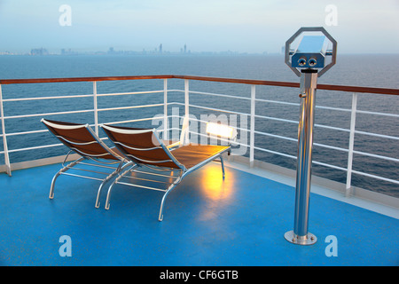 Zwei beleuchtete Liegestuhl und Fernglas auf Schiff mit Blick auf die Stadt am Abend Stockfoto