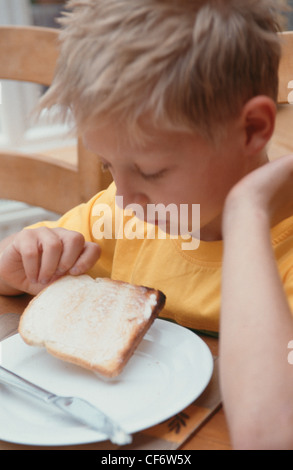 Männliches Kind stachelige blonde Haare tragen gelbe und grüne t-Shirt sitzt am Holztisch auf der Suche nach unten halten Stück mit Butter Stockfoto