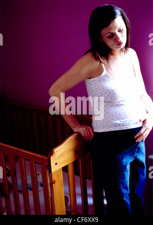 Hochschwangere Frauen lange brünette Haare tragen florale Weste Top und Jeans lehnte sich gegen Ende des hölzernen Kinderbett berühren Stockfoto