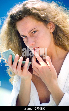 Weibliche langen lockigen dunkel blonde Haare trägt weißen Bademantel hält grün bilden kompakte, Lipgloss auf die Lippen auftragen Stockfoto