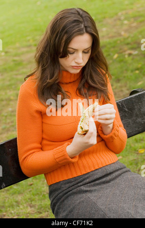 Weibchen tragen orange Rollkragen Pullover und grauen Rock sitzt auf der Parkbank Sandwich zu essen Stockfoto