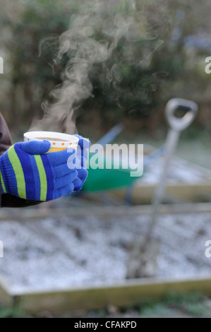 Lady Gärtner Hände in Handschuhe halten dampfenden Tasse Kaffee im Garten, mit frostigen Garten im Hintergrund, Januar Stockfoto