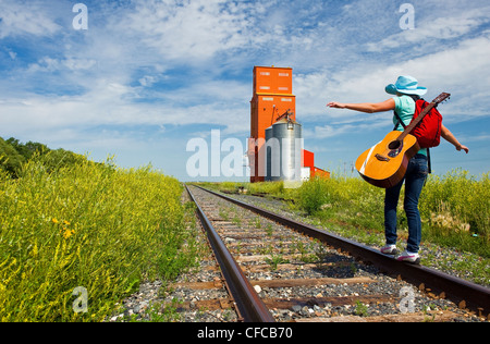 18 Jahre altes Mädchen mit Gitarre zu Fuß entlang der Eisenbahn mit verlassenen Getreidesilo im Hintergrund, Carey, Manitoba, Kanada Stockfoto