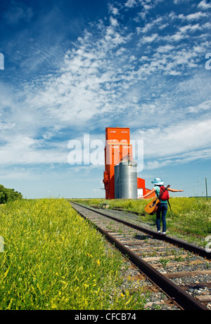 18 Jahre altes Mädchen mit Gitarre zu Fuß entlang der Eisenbahn mit verlassenen Getreidesilo im Hintergrund, Carey, Manitoba, Kanada Stockfoto