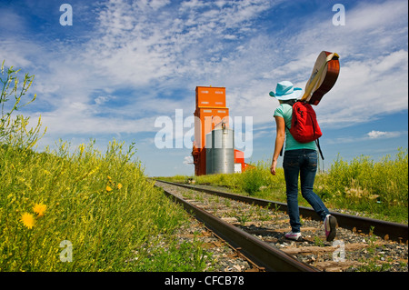 18 Jahre altes Mädchen mit Gitarre zu Fuß entlang der Eisenbahn mit verlassenen Getreidesilo im Hintergrund, Carey, Manitoba, Kanada Stockfoto