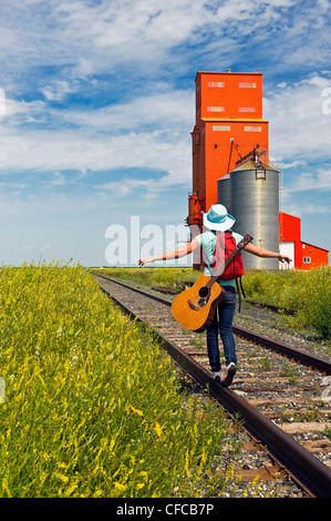 18 Jahre altes Mädchen mit Gitarre zu Fuß entlang der Eisenbahn mit verlassenen Getreidesilo im Hintergrund, Carey, Manitoba, Kanada Stockfoto