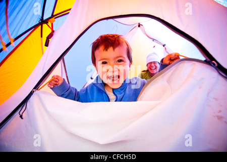 ein kleiner Junge öffnet und schließt die Zelttür mit Reißverschluss, während er auf einen blauen Schlafsack steht. Das Zelt ist Orange und blau. Die Zertifizierungsstelle Stockfoto