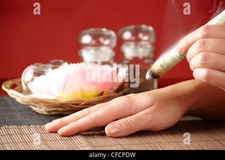 TCM-Traditionelle chinesische Medizin. Hand, die Anwendung von Moxa Stick Therapie, natürlicher Kräutern in Gläsern im Hintergrund Stockfoto