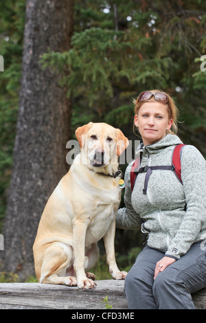 Porträt von Frau Wanderer und ihre gelben Labrador Retriever Hund, sitzend auf einem umgestürzten Baumstamm. Banff Nationalpark, Alberta, Kanada. Stockfoto