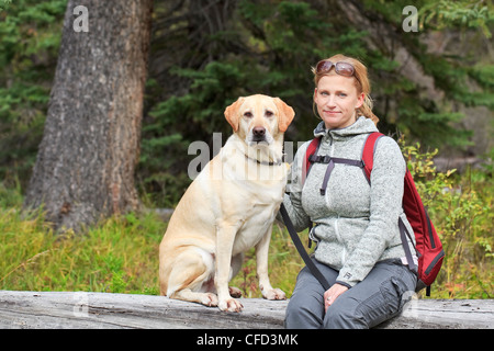Porträt von Frau Wanderer und ihre gelben Labrador Retriever Hund, sitzend auf einem umgestürzten Baumstamm. Banff Nationalpark, Alberta, Kanada. Stockfoto