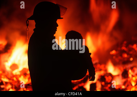 Silhouette der Feuerwehrmann mit Kind mit brennenden Haus darüber hinaus, British Columbia, Kanada. Stockfoto