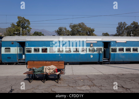 ein Mann schläft auf einer Bank am Bahnhof Kalka im indischen Bundesstaat Haryana, mit blauen Eisenbahnwaggons Stockfoto