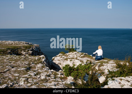 Frau sitzt auf Felsen im Hoegklint Naturreservat und bewundert Küste, Visby, Gotland, Schweden Stockfoto