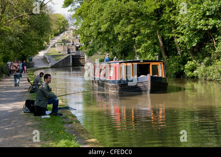 15-04 fährt auf malerischen Wasserstraße (fünf Aufstieg Schlösser, Leeds Liverpool Canal - Paar an Bord) Angler und Wanderer - Bingley, W Yorkshire, England, UK. Stockfoto