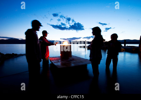 Eine Gruppe von vier jungen Erwachsenen stehen um einen schwimmenden Feuer bei Sonnenuntergang an einem See in Idaho. Stockfoto