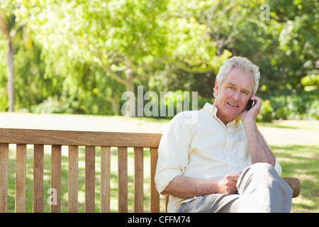 Lächelnd und Blick voraus, als er einen Anruf beim Sitzen auf einer Bank macht Mann Stockfoto