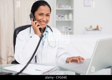 Halten Sie ihr Telefon auf ihrem Laptop tätiger Arzt lächelnd Stockfoto