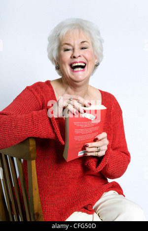 Ältere Dame in helle Kleidung, Lächeln und setzte sich auf den Stuhl mit Buch in der Hand gegen einen weißen Hintergrund Stockfoto