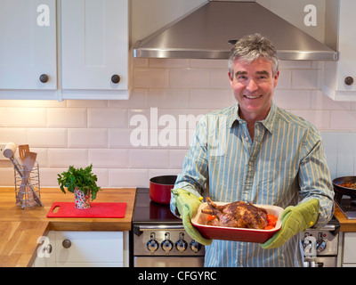 Mann KOCHEN HUHN Lächeln Entspannt zuversichtlich reifer Mann in der zeitgenössischen Küche bietet eine warme, frisch gekochte Mahlzeit von Hähnchen und Gemüse Stockfoto