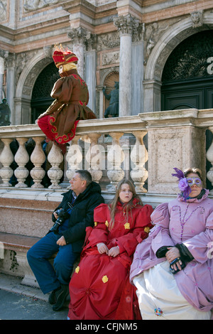 Familie im Karnevalskostüm sitzen auf einer Bank schauen gelangweilt als Karneval Charakter Posen auf einer Wand in Ihrer Nähe St. Marks Platz Venedig Stockfoto