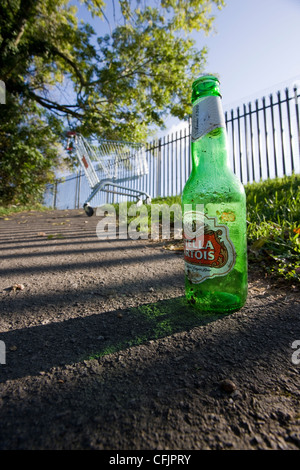 Leere Bierflasche der Marke Stella Artois auf dem Bürgersteig mit einem verlassenen Einkaufswagen im Hintergrund Stockfoto